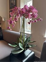 Full view of artificial orchid plant showing cluster of pink spotted flowers and broad green leaves in black ceramic planter.