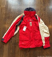 Front flat lay image of a red Helly Hansen ski jacket with white sleeves and black detailing on a wooden floor.