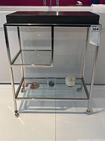 Frontal view of modern end table showing slate-colored top, chrome frame, glass shelves, and caster wheels.