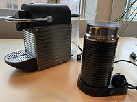 Photo of side view of the black and chrome espresso machine with the milk frother alongside it on a table.