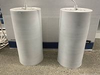 Two white cylindrical pendant lamps shown side by side standing on concrete floor, showing cylindrical shape and paint condition.