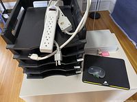 Lot displayed showing black plastic stacking filing trays, power bars, fireproof cashbox, mouse, and mousepad on a table.