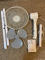 Disassembled fan parts laid out including fan head with grille, three fan blades, motor housing, vertical pedestal metal pieces, and instruction manual on carpet.