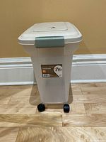 Full view of the translucent rectangular pet food container with lid and castors on a hardwood floor and beige wall background.