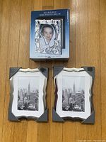 Photo shows the silver-plated baby photo album in original box and two chrome photo frames with wavy edges still partially covered with protective triangles containing black and white photo print of city skyline.