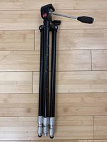 Folded black metal tripod with silver-colored extendable leg sections and a black tilting head with a handle, viewed from above on a light wood floor.
