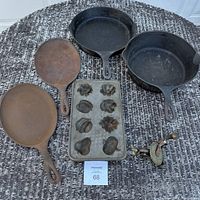 Top-down view of four cast iron skillets and pans, vintage cast iron baking mold with 9 shaped cavities, and hand crank metal grinder on woven surface.