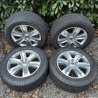 Four chrome alloy wheels with tires laid out on gravel, visible wear on tires