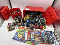 Photo showing 9.75lb box of assorted Lego bricks, several Lego instruction booklets, and two large red Lego storage bins partially open with Lego bricks spilling out.
