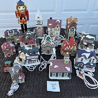Full assortment of vintage ceramic holiday village houses grouped together on the ground with electric cords visible and a wooden nutcracker soldier figure behind the houses.