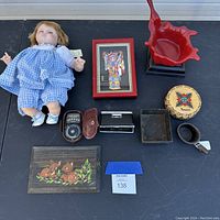 Overview of all items showing porcelain doll, framed artwork, red sculpture, round wooden box, wooden plaque with fawns, exposure meter, metal item, velvet tray, and wooden cup.