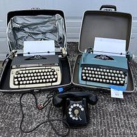 Two suitcase style mechanical typewriters with paper loaded, one beige Smith-Corona Classic 12 and one blue Hermes 3000, alongside a black vintage rotary dial telephone