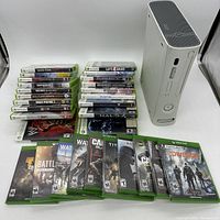 Photo showing white Xbox 360 console (no cords) standing vertically with stacks of Xbox 360 and Xbox One video games arranged beside it.