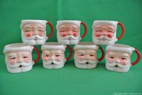 Front view of 7 Santa Claus cups arranged in two rows showing 4 with eyes open and 3 with eyes closed, placed on green background.