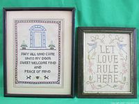 Front view of two framed cross stitch pieces, one larger with a welcoming phrase and decorative border, the other smaller with 'LET LOVE RULE HERE' phrase and floral design.