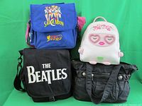 Four bags displayed against green cloth background: blue Sailor Moon backpack, white Betsey Johnson backpack, black Beatles shoulder bag, black Kenneth Cole tote bag.