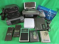 Photo showing multiple vintage electronic audio devices including nightstand radios, cassette walkmans, and recorders arranged on green background.