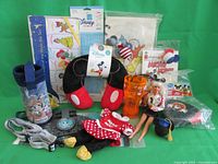 Display of assorted Disney items including bib set, stencil, pillowcase, plush ears, Barbie doll, cups, keychains, and badges.