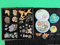 Overview of the jewelry pieces including pins, brooches, earrings, and trinket boxes on black cloth surface.