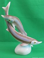 Front view of one porcelain sturgeon sculpture with a visible broken fin towards the bottom of the fish near the base, showing detailed body and fins, against green backdrop.