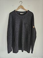 Full front view of the dark gray Fjallraven crewneck sweater on a hanger, showing its overall design and texture.
