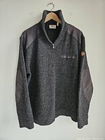 Full view of the men's Fjallraven dark gray half zip sweater hanging on a hanger showing the front with reinforced shoulders and elbow patches.