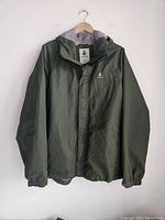 Front view of green waterproof jacket with hood, zipper, and button closure, hanging on wooden hanger.