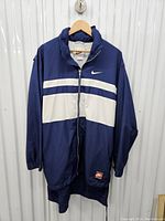 Full front view of the vintage Nike navy blue and white jacket on a hanger showing front zipper, white stripes, Nike swoosh, and Nike patch on bottom front.