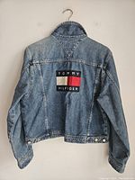 Back view of vintage Tommy Hilfiger denim jacket showing large logo patch with text 'Tommy Hilfiger' in black, white, red, and navy.