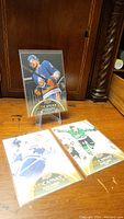Three hockey cards displayed on a wooden surface: a card featuring an Edmonton Oilers player in blue and orange uniform on a stand, and two cards lying flat showing players from the Toronto Maple Leafs and Dallas Stars.
