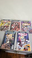 Photo showing five Harley Quinn comic books laid out flat, colorful covers visible displaying different Harley Quinn poses and themes.