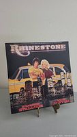 Front cover of Rhinestone vinyl record featuring Sylvester Stallone and Dolly Parton standing in front of yellow taxi and city skyline at night.