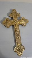 Front angle view of gold-tone metal crucifix cross mounted on a wall, showing detailed relief of Jesus Christ and decorative ends of the cross.