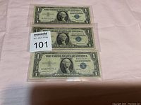 Three $1 Silver Certificate bills laid out vertically on a light-colored fabric surface in clear protective sleeves