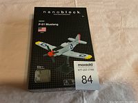 Closed box of Nanoblock P-51 Mustang micro-sized building block kit shown on beige fabric background with MaxSold lot card.