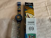 Blue Rugrats movie themed wristwatch alongside its colorful original box, placed on a white cloth background. The watch has character art on the wristband and face, with the box showing a character from the movie.