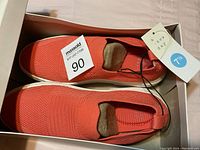 Top-down view of coral knit sneakers inside shoebox with size tag showing 7½ and 'A New Day' branding, soles white and clean.