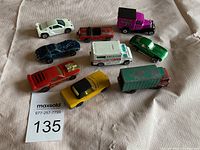Nine die-cast toy vehicles arranged on a fabric surface including sports cars, trucks, and vans showing signs of heavy use and paint chipping.
