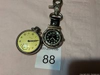 Two watches placed on a cloth background, showing the face and features of both watches including the pocket watch and the keychain clip watch.