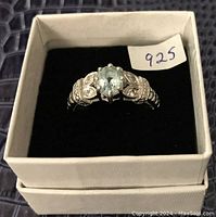 Front view of sterling silver ring showing carved band with accent stones and central light blue faceted gemstone in box with '925' label.