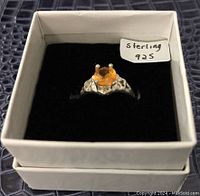 Front view of sterling silver ring with orange round gemstone in prong setting inside white box with black interior.
