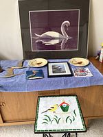 Full lot on wooden cabinet, showing large framed swan print, two small bird plates, four square pieces (two ceramic bird tiles, two brass flying geese), metal hummingbird figure, framed duck picture, and mosaic bird table.