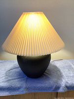 View of the lamp illuminated, highlighting the cream accordion pleated shade and wide black ceramic base.