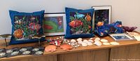 Two blue throw pillows with bright fish illustrations, four metal shell-shaped drawer pulls, assorted seashells including orange scallop and white clam shells, framed fish prints, and blue wooden stool on display.