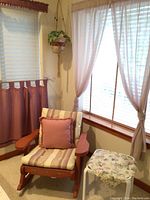 Full seating area showing rocking chair with cushions, macrame hanging faux plant, mauve and beige curtains with valances and rods, sheer curtains, small floral cushioned side table.