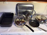 Set of stainless cookware on white cloth showing pans, lids, teapot, broiler pan, and roasting pan.