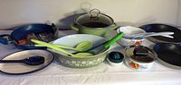 Full view of all cookware pieces including a navy blue Rachel Ray skillet, olive green enamel pot, Pyrex casserole, white floral metal pots, Farberware skillets, and plastic utensils