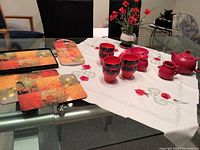 Photo showing red ceramic tea set pieces arranged on embroidered square white linen tablecloth with red poppy designs. Also showing some floral arrangement and part of place mats with floral pattern.