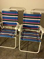 Pair of striped beach chairs with blue, green, orange, and white stripes, metal white frames and armrests, foldable design visible on concrete floor.