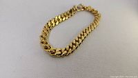 Gold-tone Cuban link bracelet with S clasp, approximately 8mm wide, laid flat on light background, multiple angles.